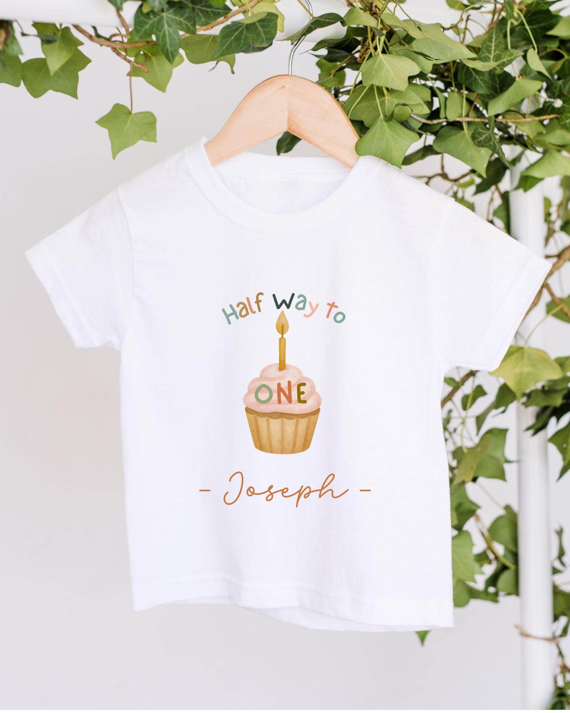 A white t-shirt with a graphic design featuring a cupcake with a candle on it, along with the text "Half Way to Joseph" printed on it.