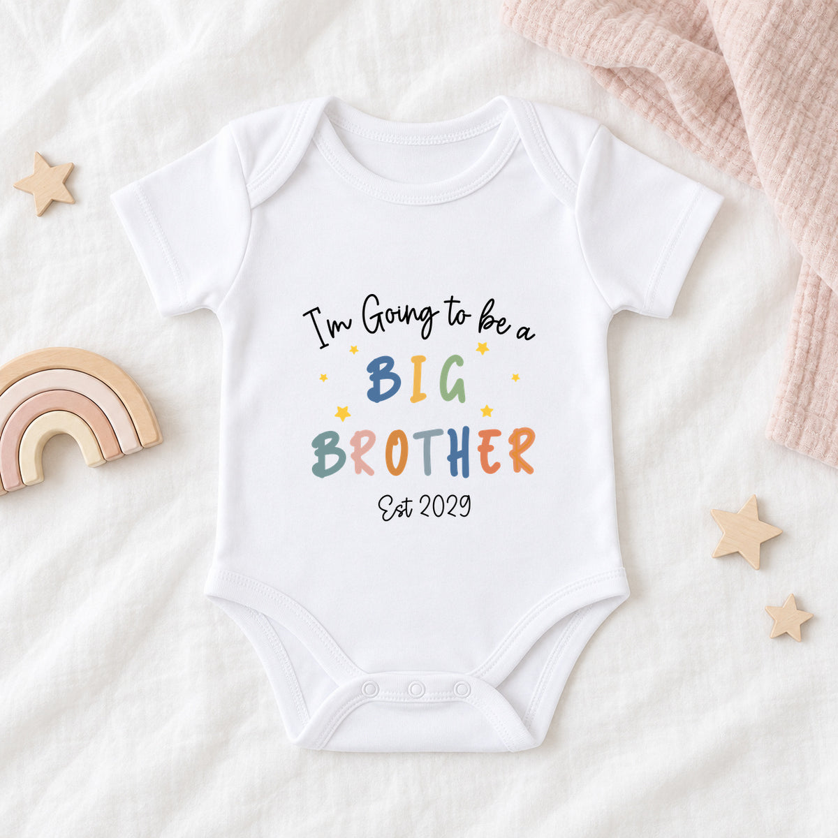 Personalised white Baby Vest with colourful I'm Going To Be A Big Brother wording and stars, styled with blush muslin and a wooden rainbow.