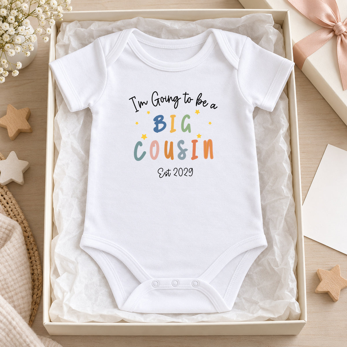White Baby Vest reading I'm Going to be a Big Cousin, Est 2029, in an ivory tissue-lined gift box beside wooden stars, a blank card and blush ribbon.