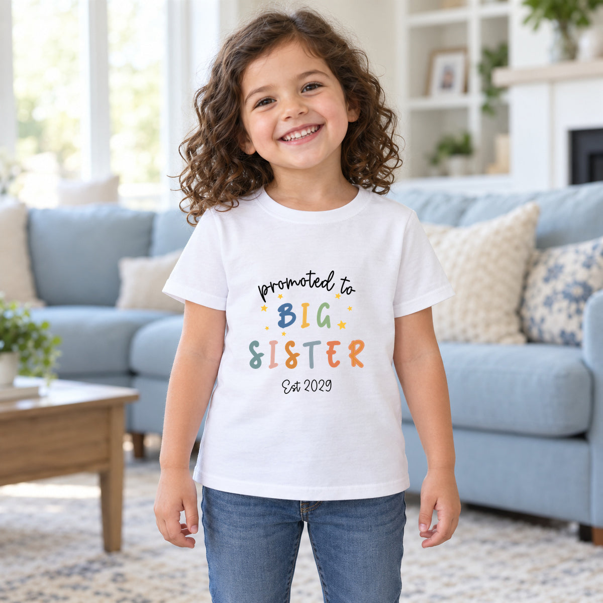 Smiling girl wearing a white Promoted to Big Sister T-shirt with colourful letters, yellow stars and Est 2029 wording, standing in front of a pale blue sofa in a bright living room.