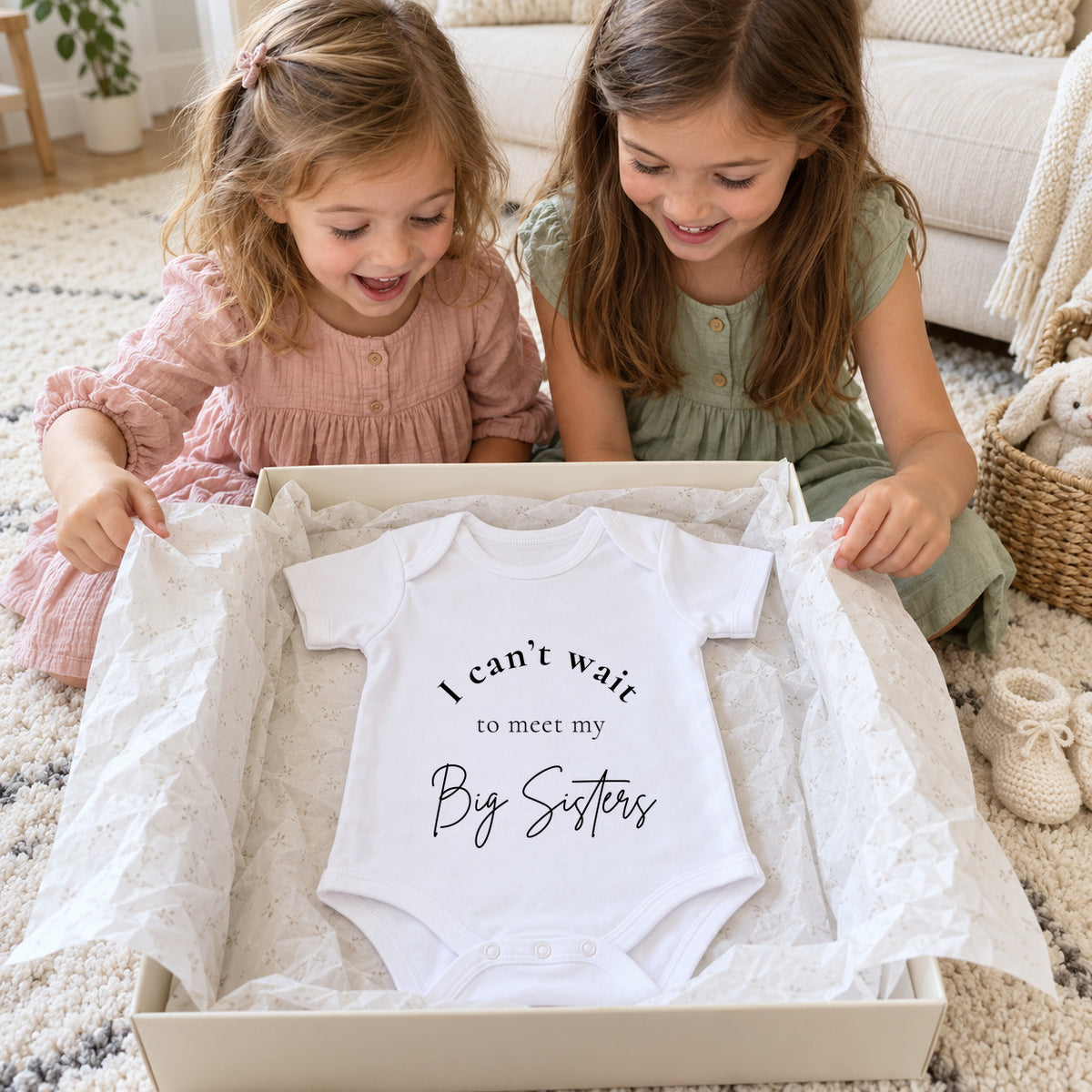 Two smiling sisters opening a cream gift box with a white Baby Vest reading I can't wait to meet my Big Sisters, on an ivory nursery rug.