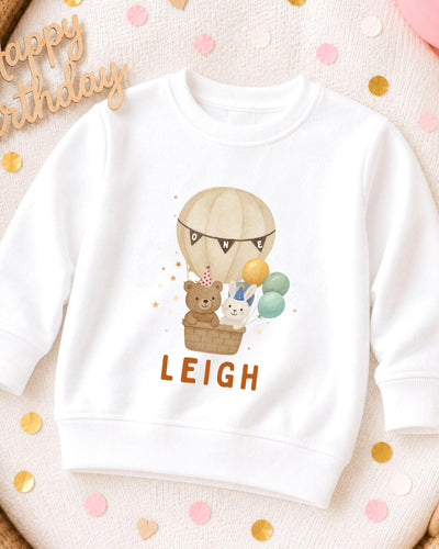 A white sweatshirt with a cartoon image of a hot air balloon carrying a teddy bear and a rabbit, along with the name "LEIGH" printed on it.