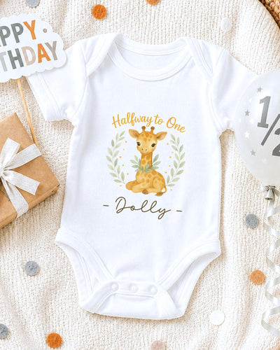 A white onesie with a giraffe design and the text "Halfway to One Dolly" printed on it, along with a "Happy Birthday" sign and confetti.