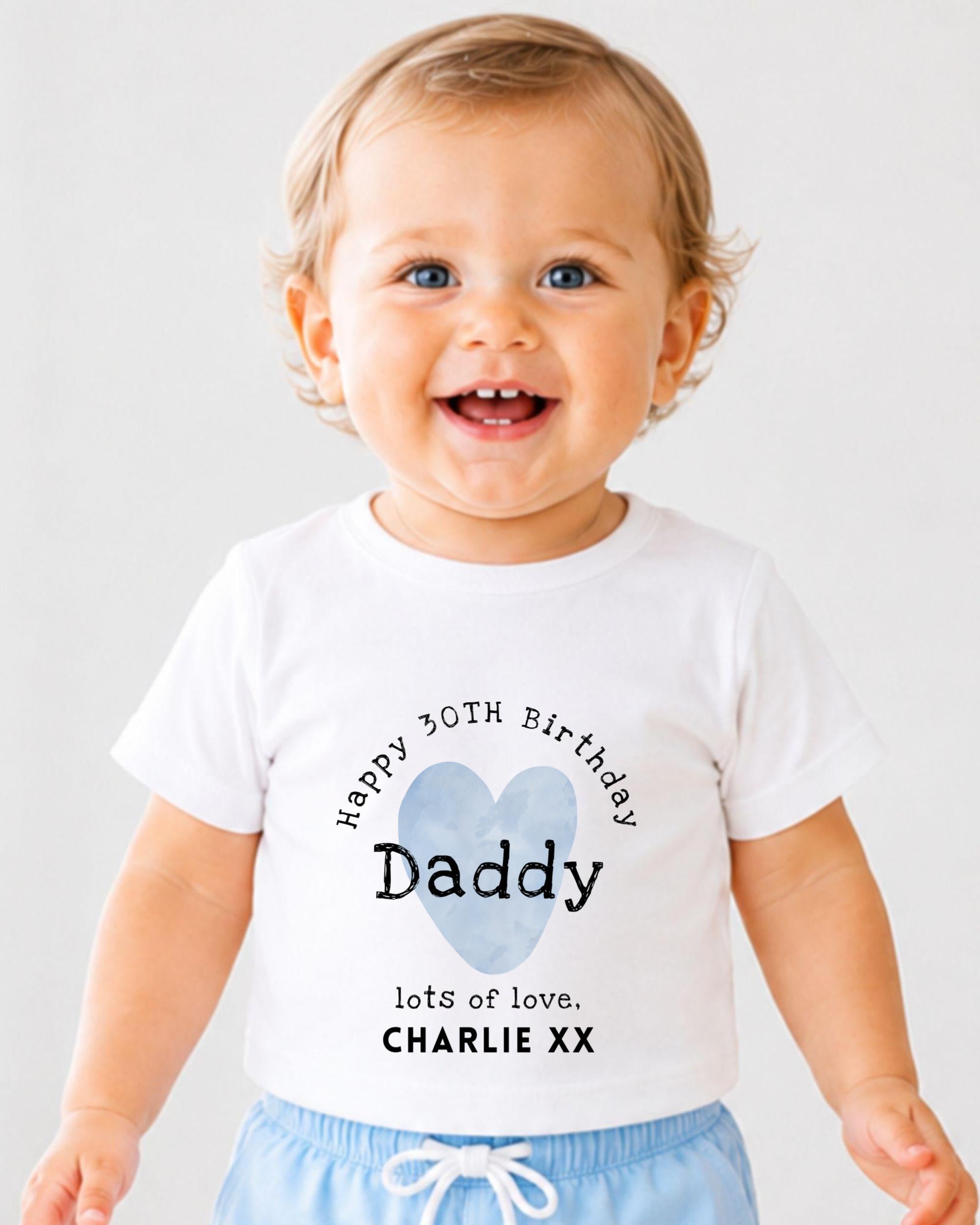 A smiling baby boy wearing a white t-shirt with a personalized message and a heart graphic.