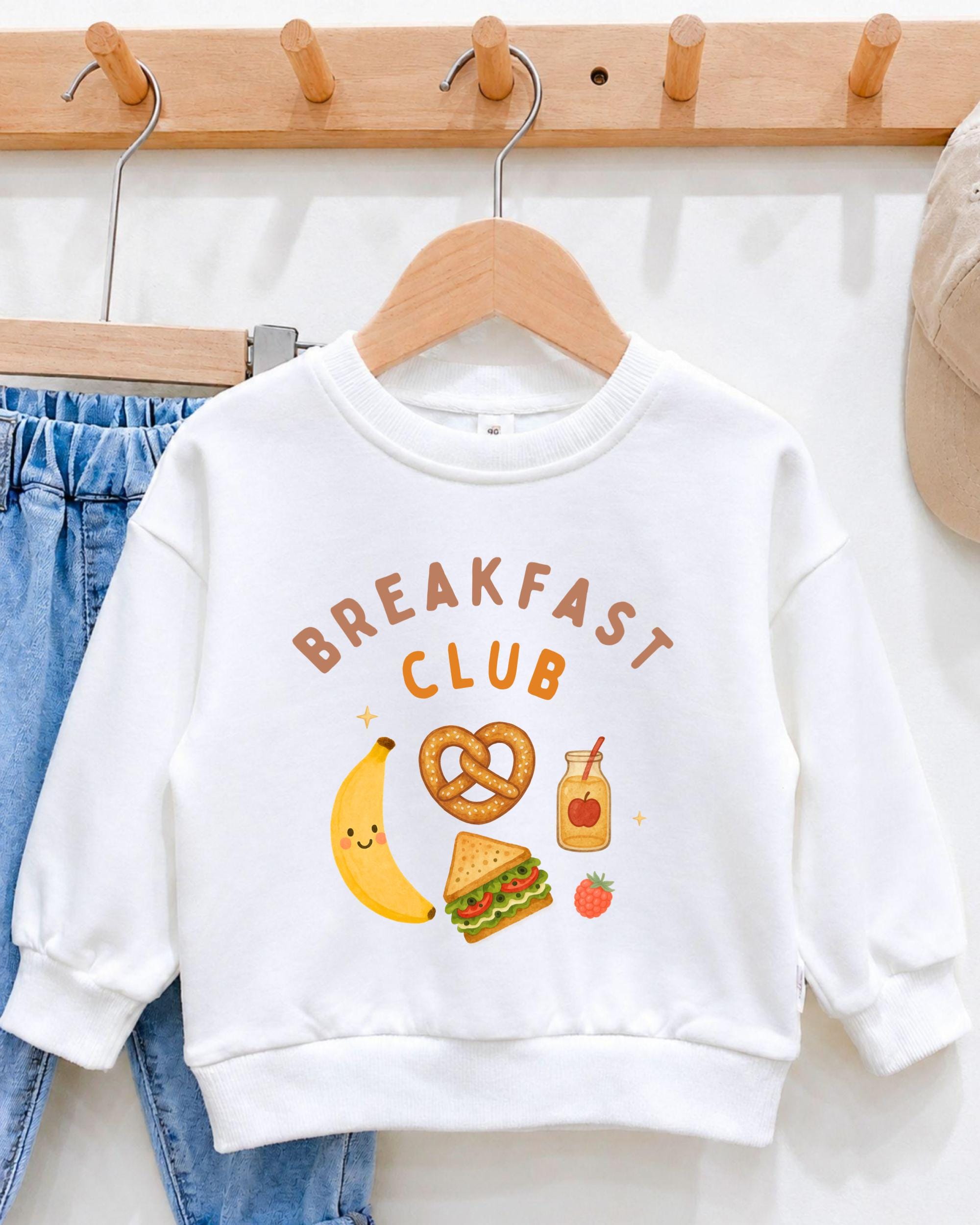 A white sweatshirt with the text "BREAKFAST CLUB" printed on it, along with various food items such as a banana, pretzel, sandwich, and apple.
