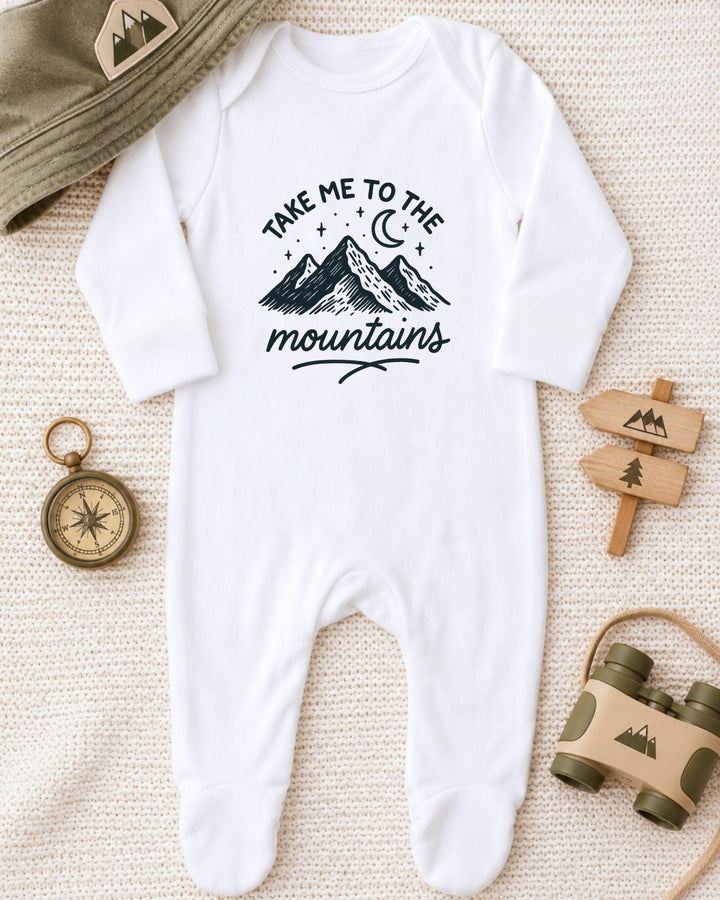A white onesie with a mountain and starry night sky design, along with a compass, a hat, and a pair of binoculars, all arranged on a beige surface.