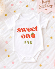 A white baby onesie with the text "Sweet on EVE" printed on it, placed on a bed with confetti and a birthday card.