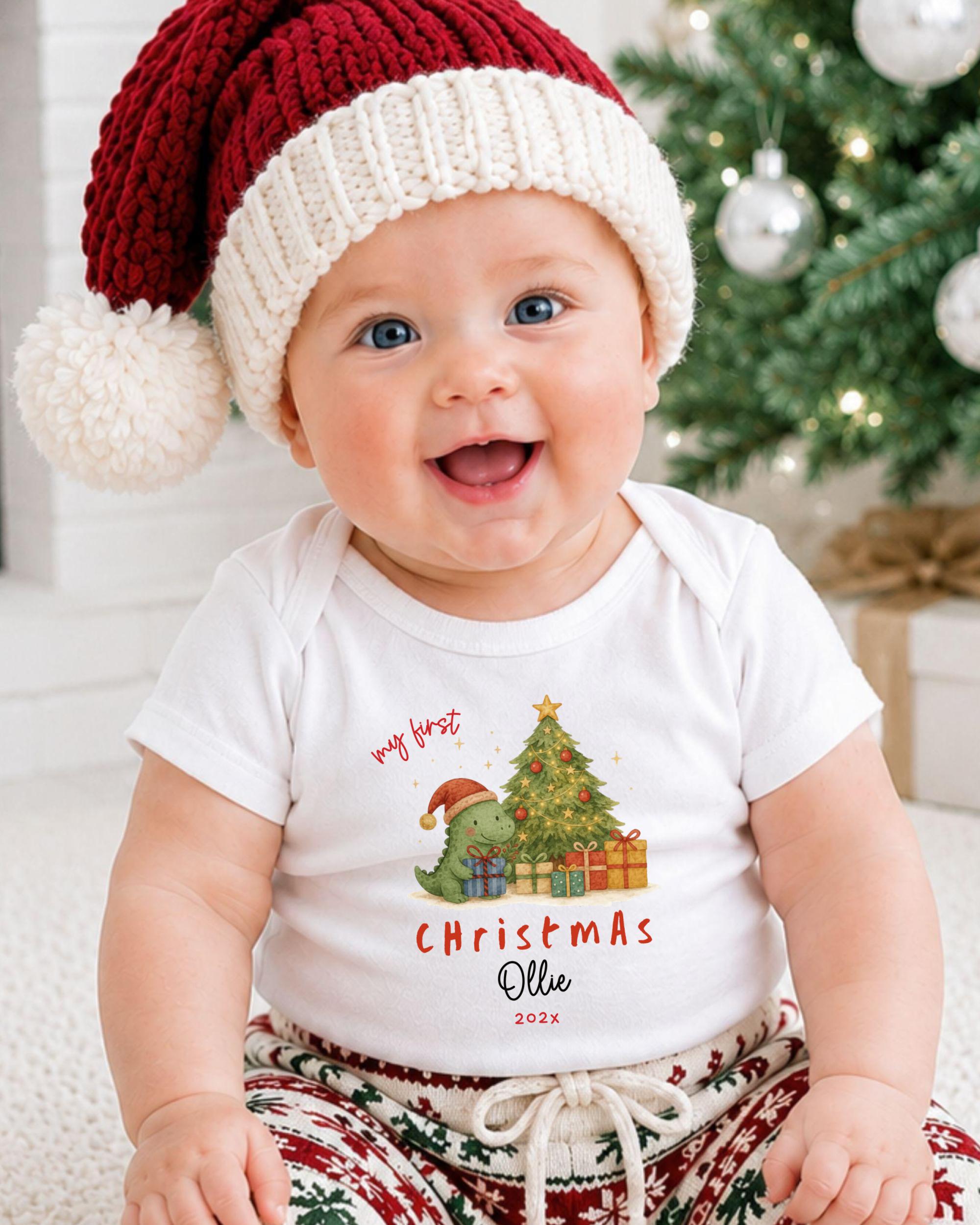 A smiling baby wearing a Santa hat and a Christmas-themed t-shirt sits in front of a Christmas tree.