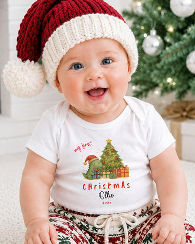 A smiling baby wearing a Santa hat and a Christmas-themed t-shirt sits in front of a Christmas tree.