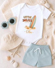 A baby's outfit, including a white onesie with a surfboard design, a beige bucket hat, and light blue shorts, along with sunglasses and seashells, are arranged on a beige surface.