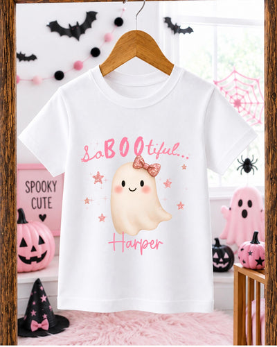 A white t-shirt with a pink ghost and the text "SaBOE LITERAL Harper" printed on it, hanging on a wooden hanger. The t-shirt is displayed in a room decorated for Halloween, with various Halloween-themed decorations and items visible in the background.