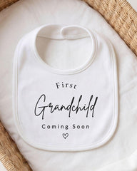A white bib with the text "First Grandchild Coming Soon" printed on it, placed on a white cloth.