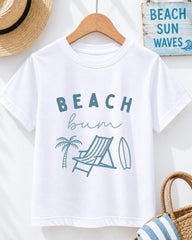 A white t-shirt with the text "BEACH butt" printed on it, along with illustrations of a beach chair, palm tree, and surfboard. The t-shirt is hanging on a wooden hanger, and there is a straw hat and a "BEACH SUN WAVES" sign in the background.