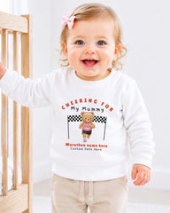 A smiling baby girl wearing a white sweatshirt with a teddy bear and text printed on it.