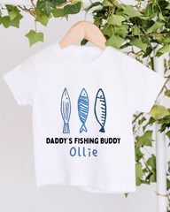 A white t-shirt with a graphic design featuring three fish and the text "DADDY'S FISHING BUDDY OLLIE" printed on it.