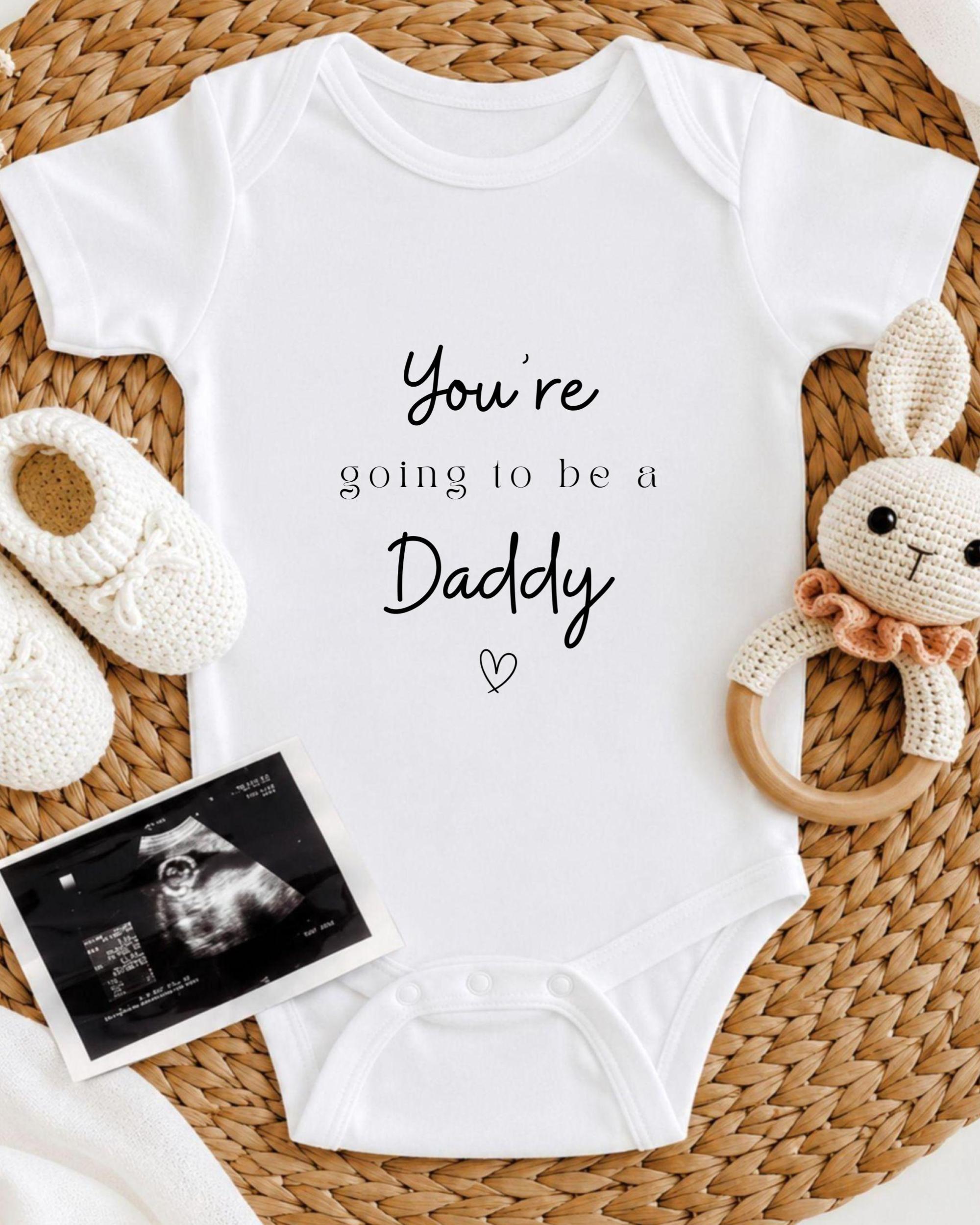 A white baby onesie with the text "You're going to be a Daddy" printed on it, along with a pair of white crocheted shoes and a stuffed bunny toy.