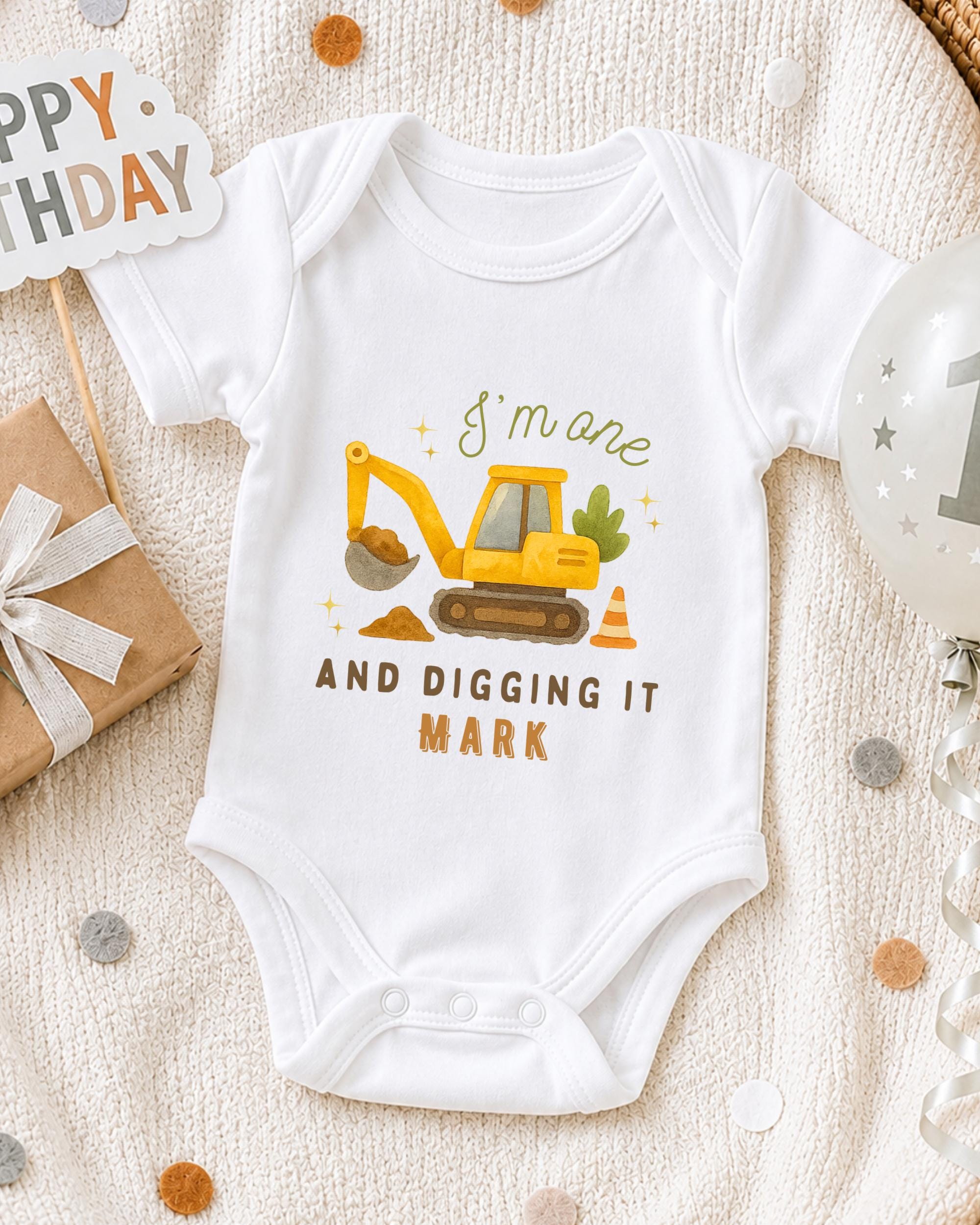 A white onesie with a yellow construction vehicle graphic and the text "I'm one AND digging it MARK" printed on it, along with a "Happy Birthday" sign and a birthday balloon.