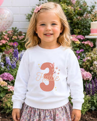 A young girl with blonde hair wearing a white sweater with the number 3 and floral designs, standing in front of a garden with colorful flowers.