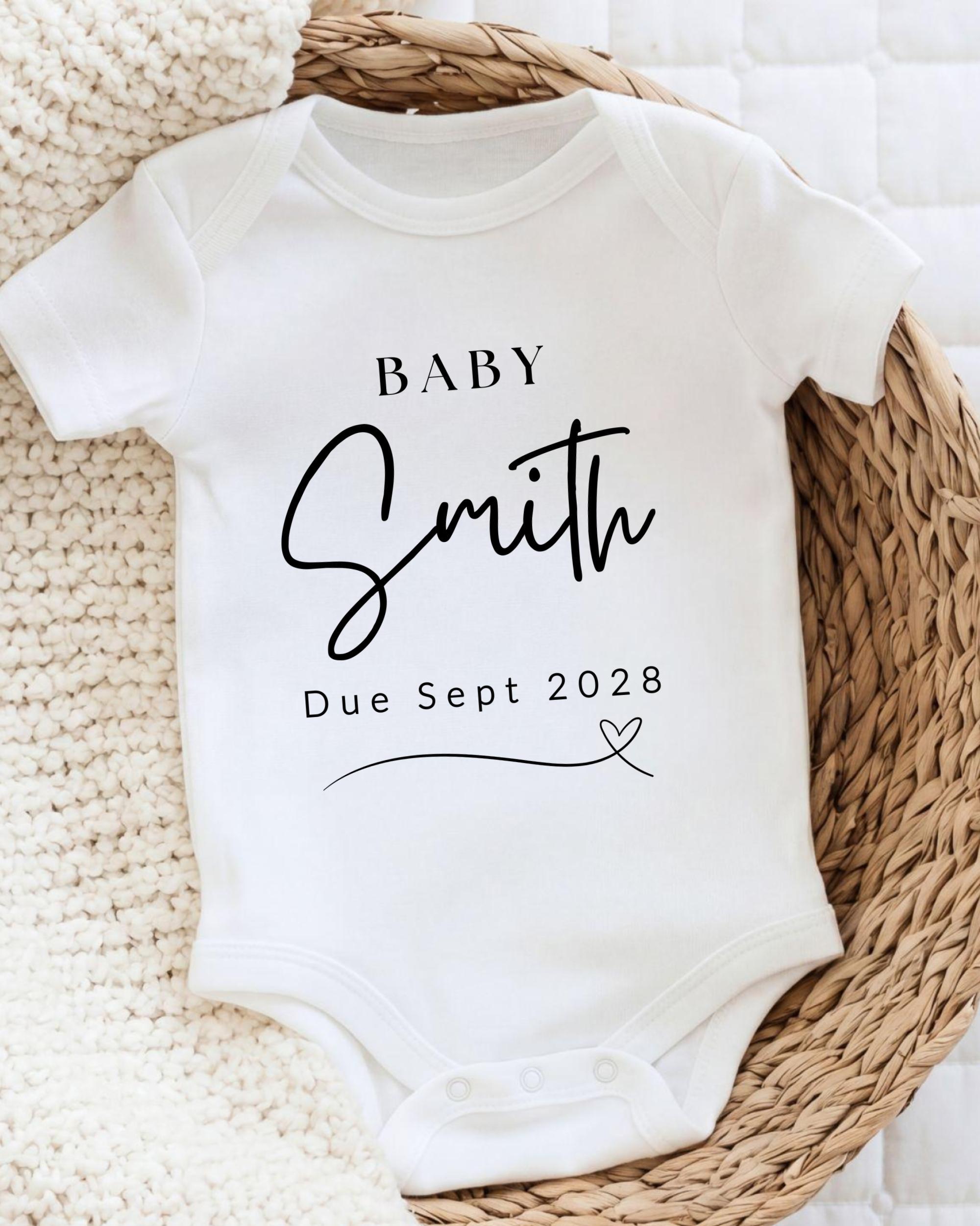 A white baby onesie with the text "BABY Smith Due Sept 2028" printed on it, placed on a woven basket.