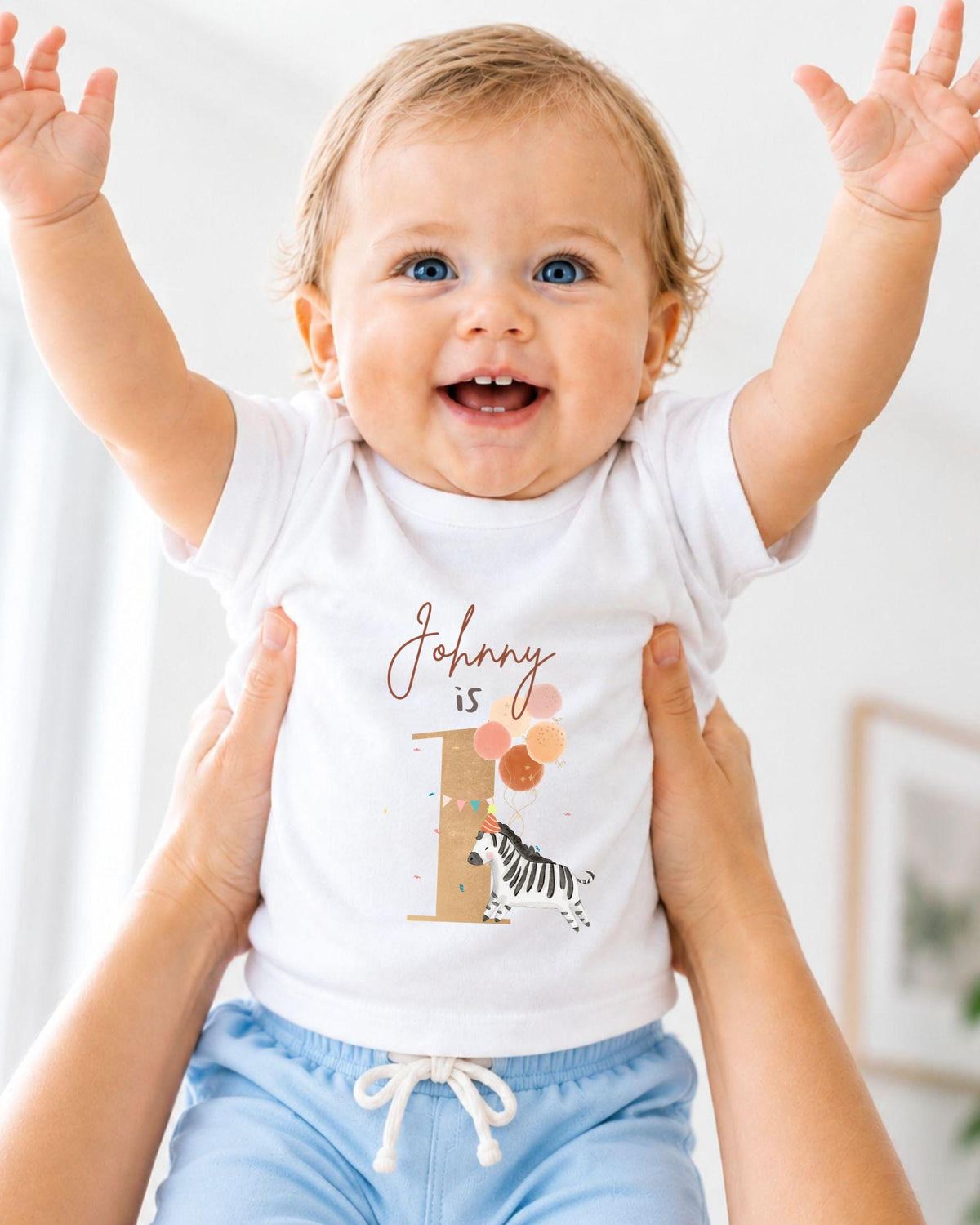 A smiling baby wearing a white t-shirt with a personalized number 1 and zebra design, being held up by someone's arms.