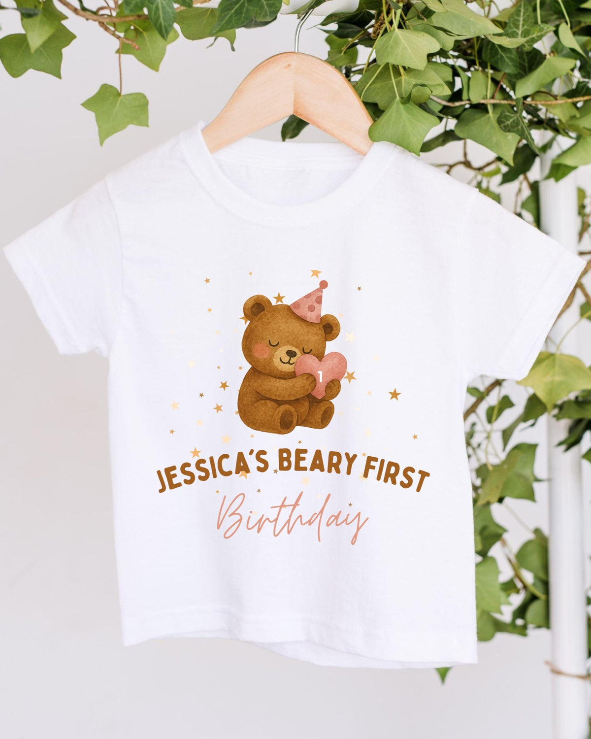 A white t-shirt with a cartoon bear wearing a party hat and holding a heart-shaped balloon, along with the text "JESSICA'S BEARY FIRST Birthday" printed on it.