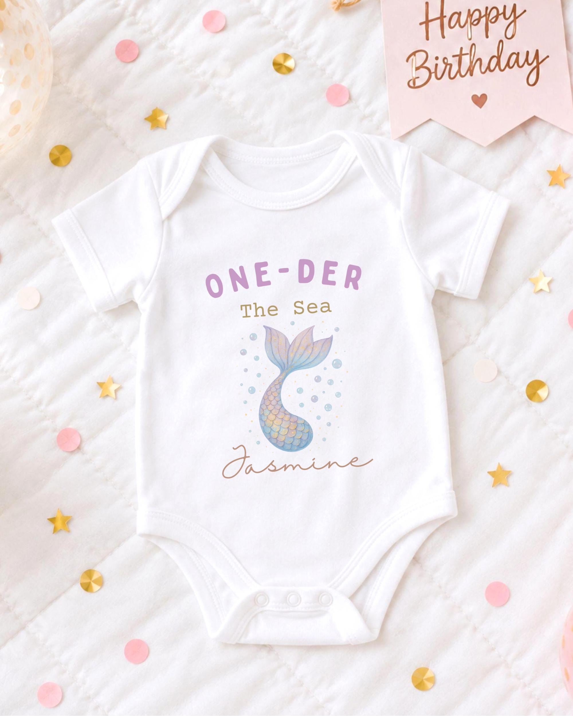 A white onesie with a mermaid design and the text "One-DER The Sea Jasmine" printed on it, placed on a bed with confetti and a "Happy Birthday" sign.