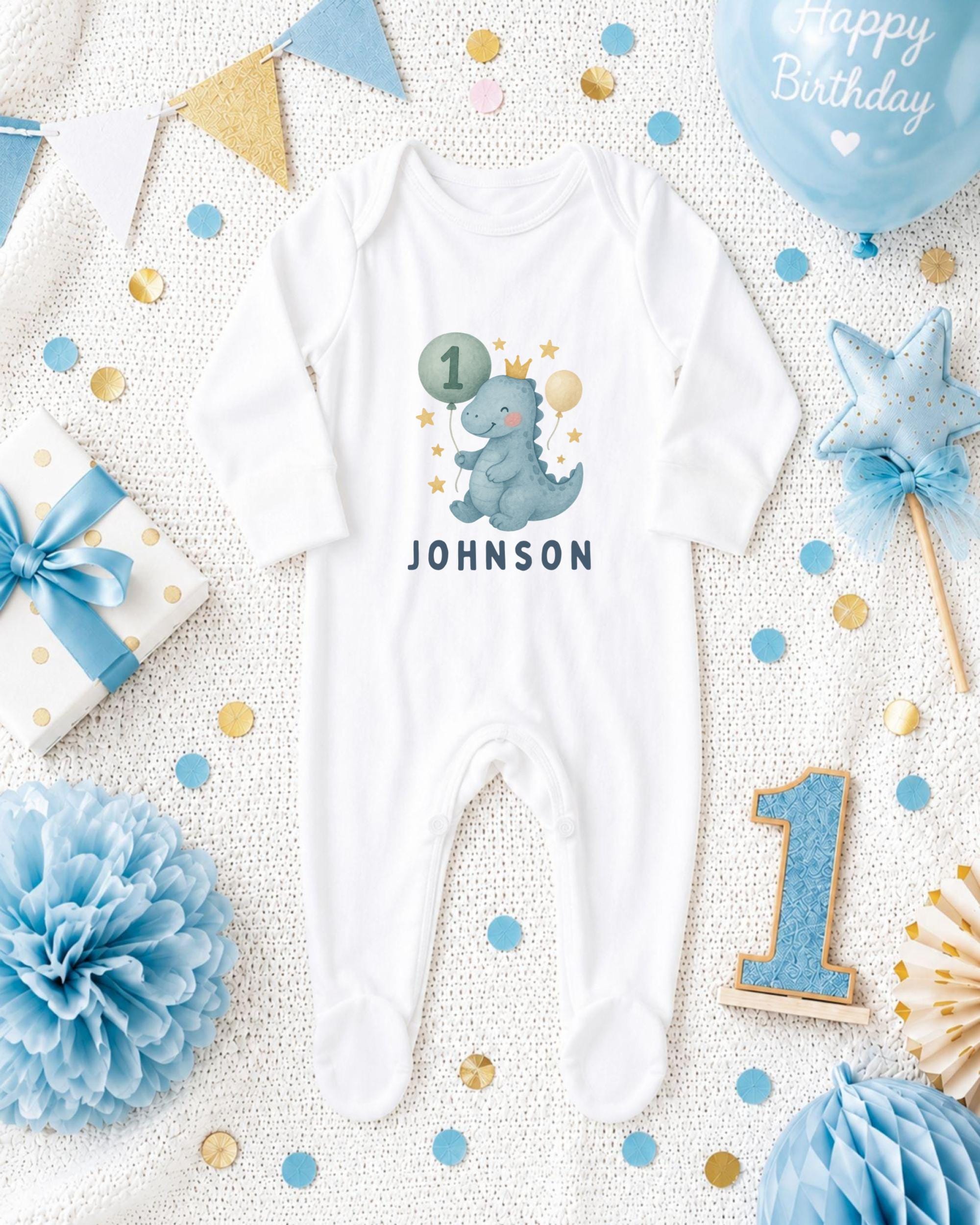 A white onesie with a dinosaur and the number 1, surrounded by birthday decorations such as a blue balloon, blue and gold confetti, and a blue and gold star.