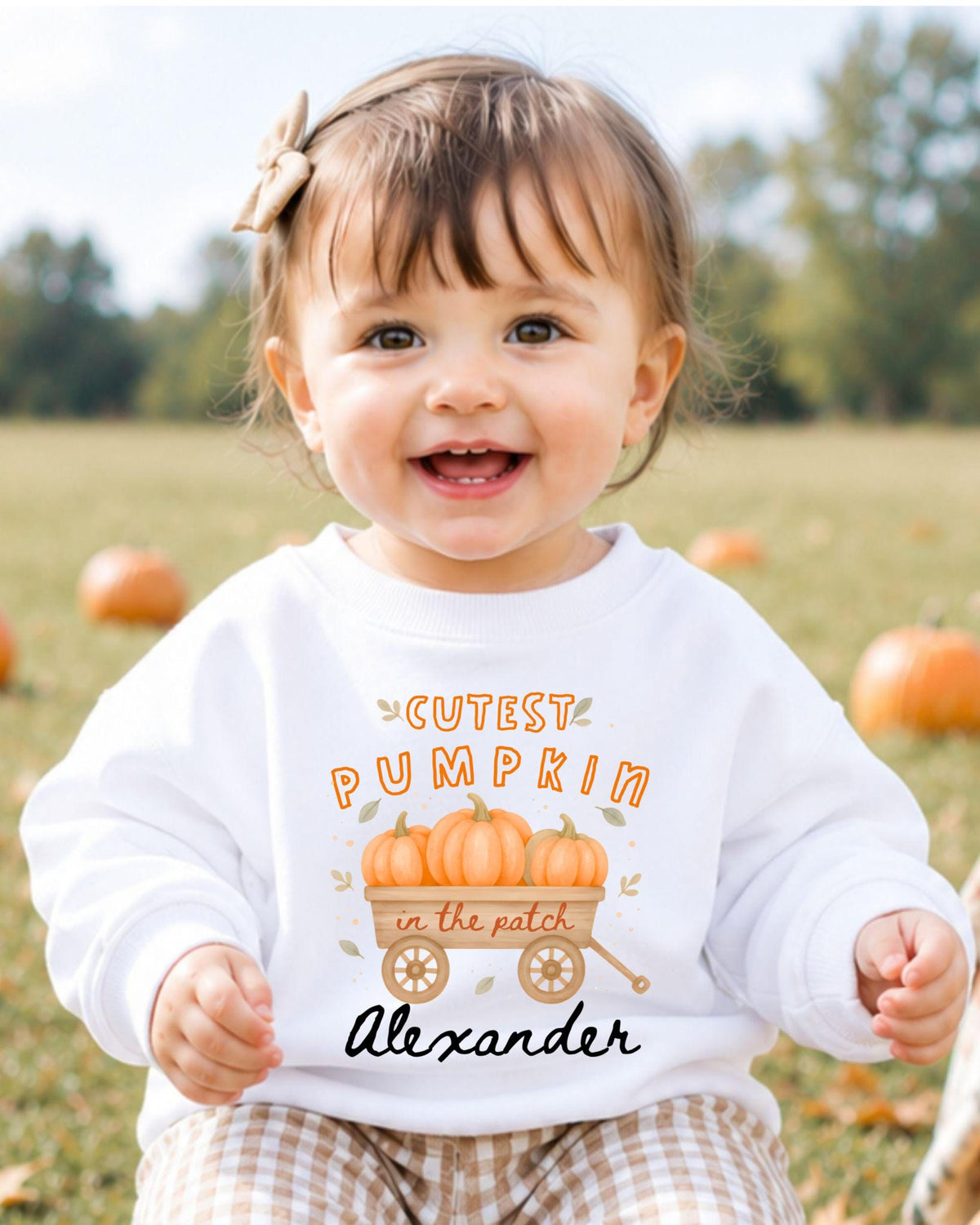 A young girl wearing a white sweatshirt with a pumpkin design and the text "CUTEST PUMPKIN in the patch Alexander" smiling at the camera.