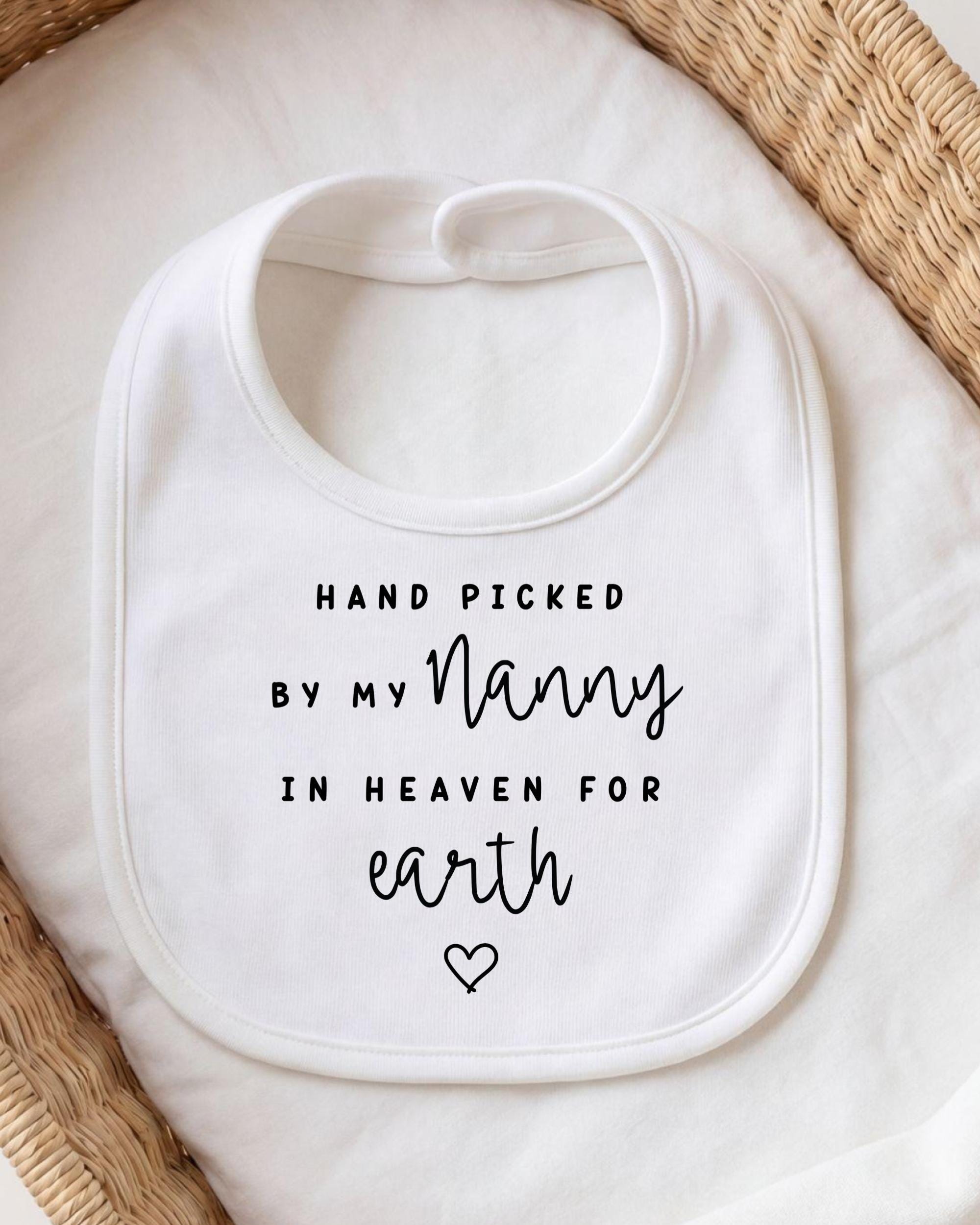 A white bib with black text that reads "HAND PICKED BY MY NANNY IN HEAVEN FOR EARTH" and a heart symbol.