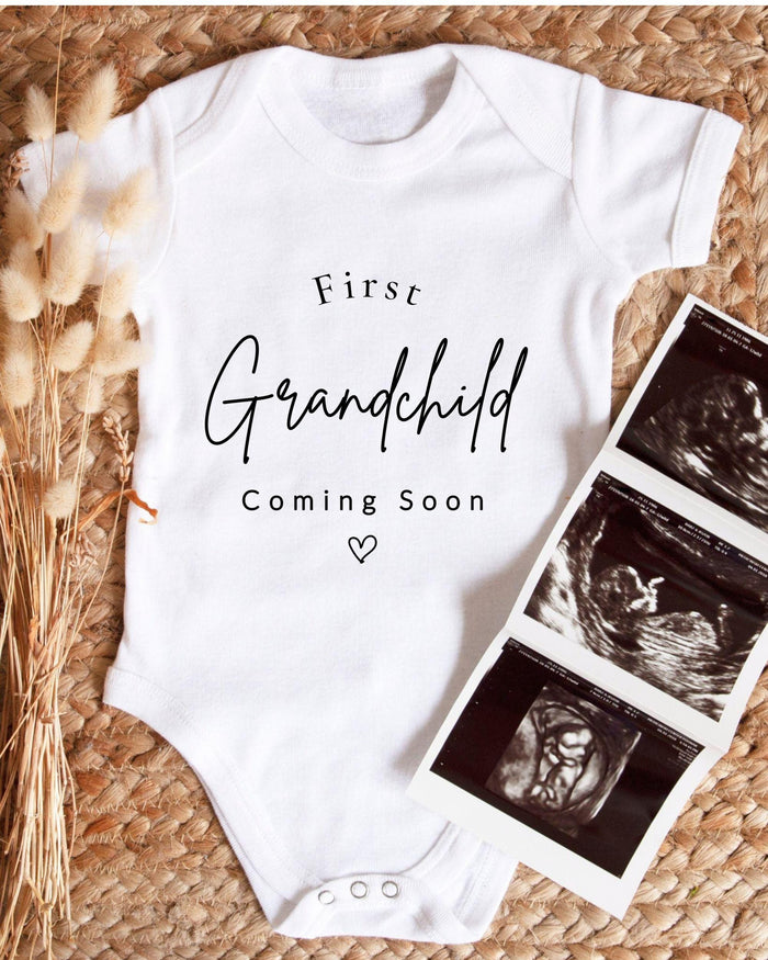 A white bodysuit with the text "First Grandchild Coming Soon" printed on it, along with a photograph of an ultrasound image of a baby's head.