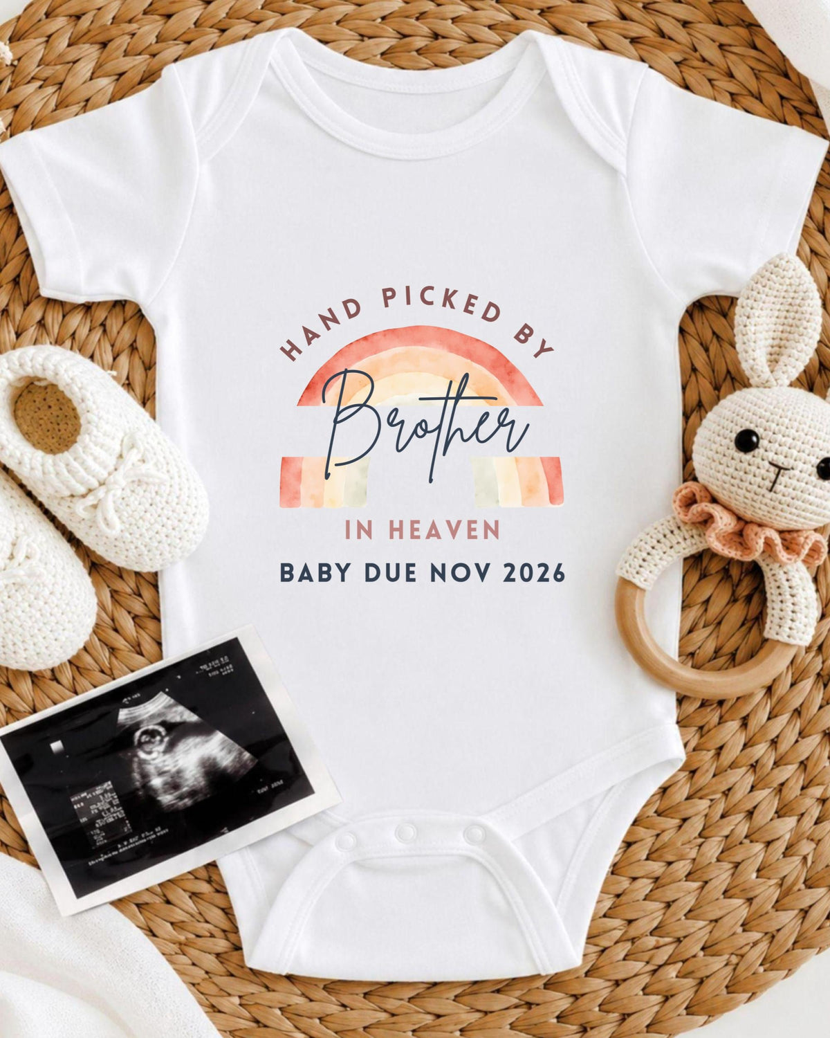 A white baby onesie with a rainbow graphic and text, along with a crocheted bunny toy and a baby's firstborn sibling announcement.