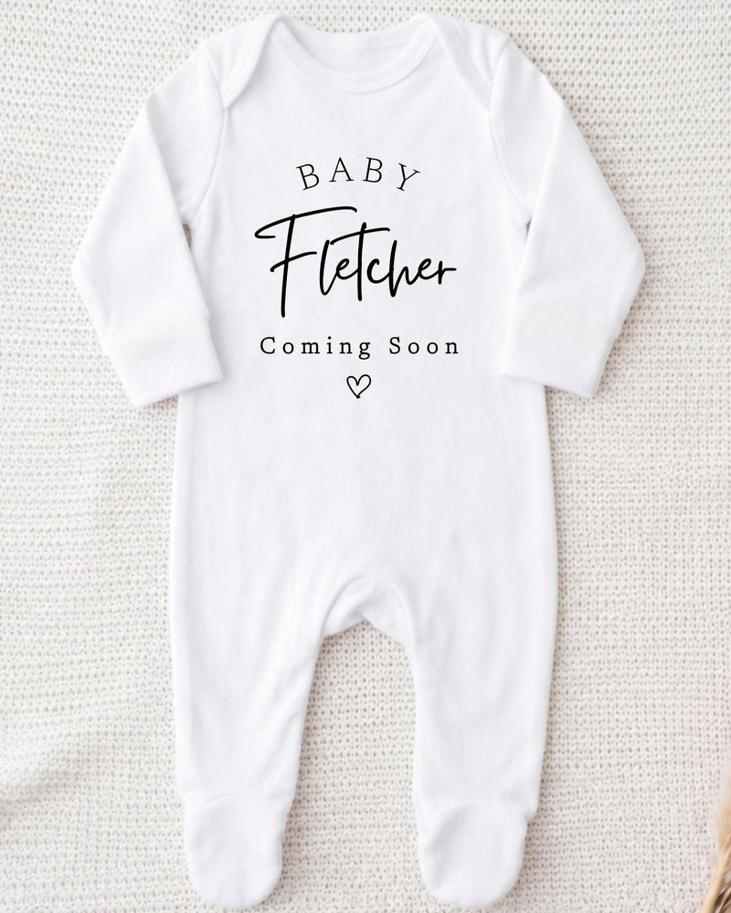 Personalisable white Baby Grow with black 'Baby Fletcher Coming Soon' serif and script lettering and a small outlined heart.