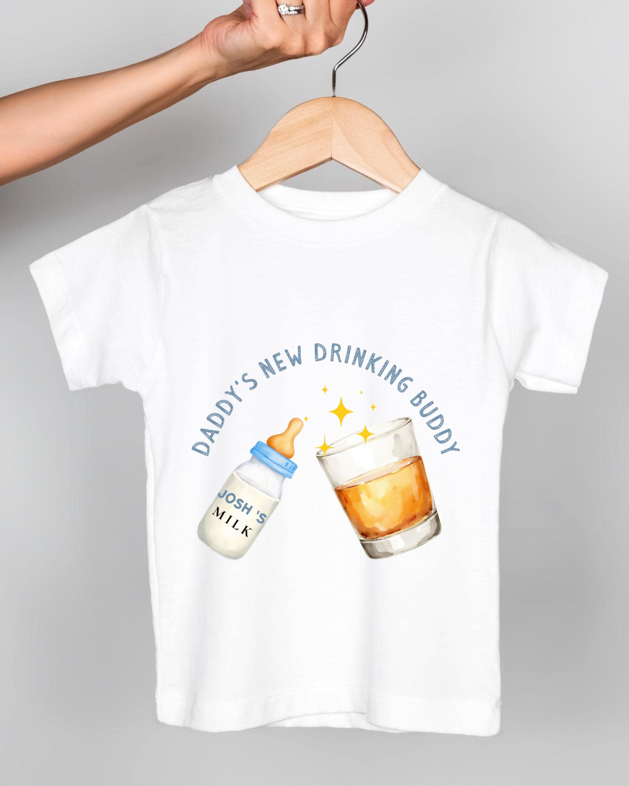 A white t-shirt with a graphic design depicting a baby bottle and a glass of amber liquid, along with the text "DADMY'S NEW DRINKING BUDDY" and a starburst effect.