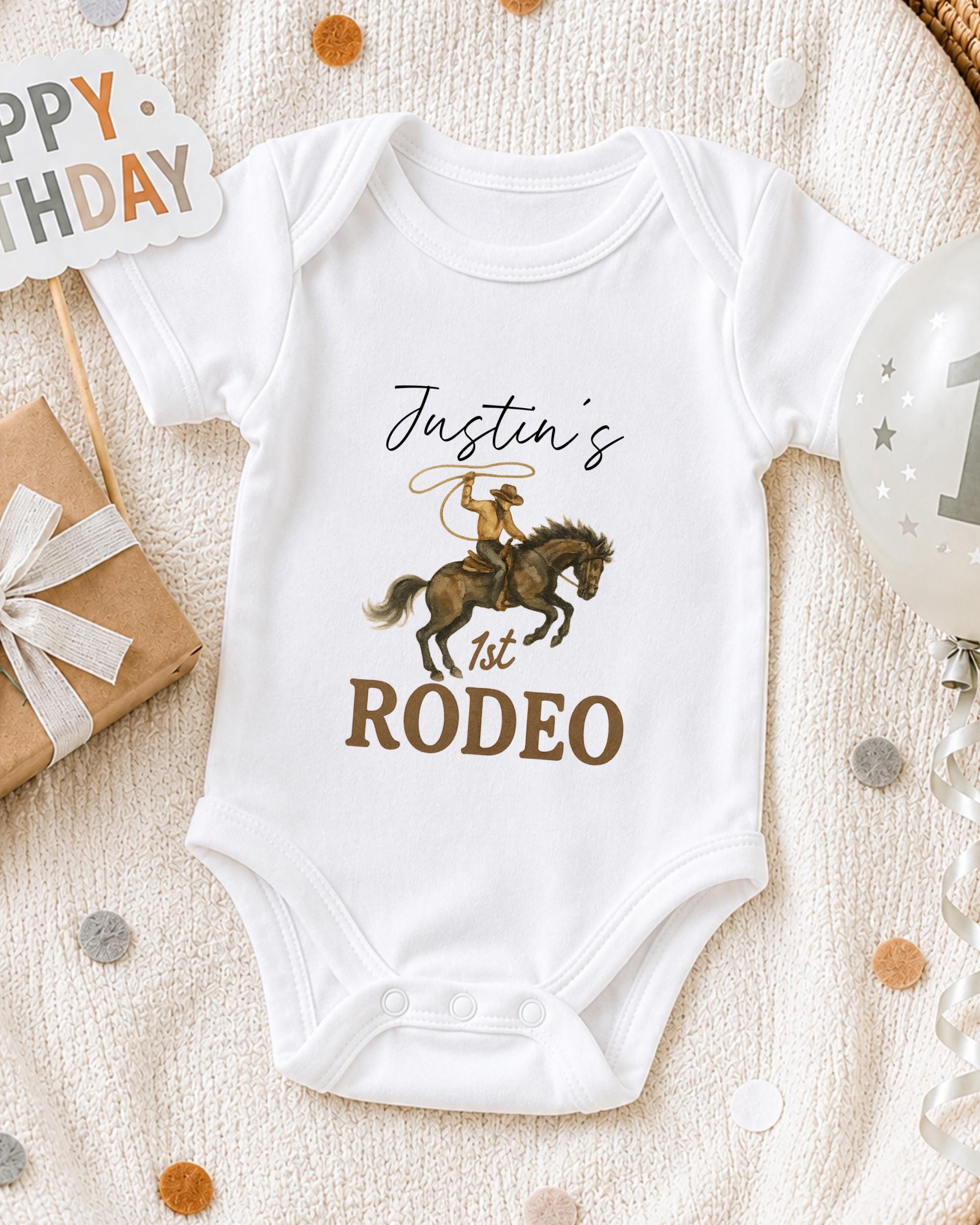 A white onesie with a cowboy graphic and the text " Justin's 1st Rodeo" printed on it, along with a birthday sign and a balloon.
