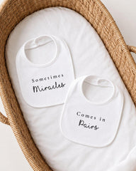 two white bibs with black text, placed in a woven basket.