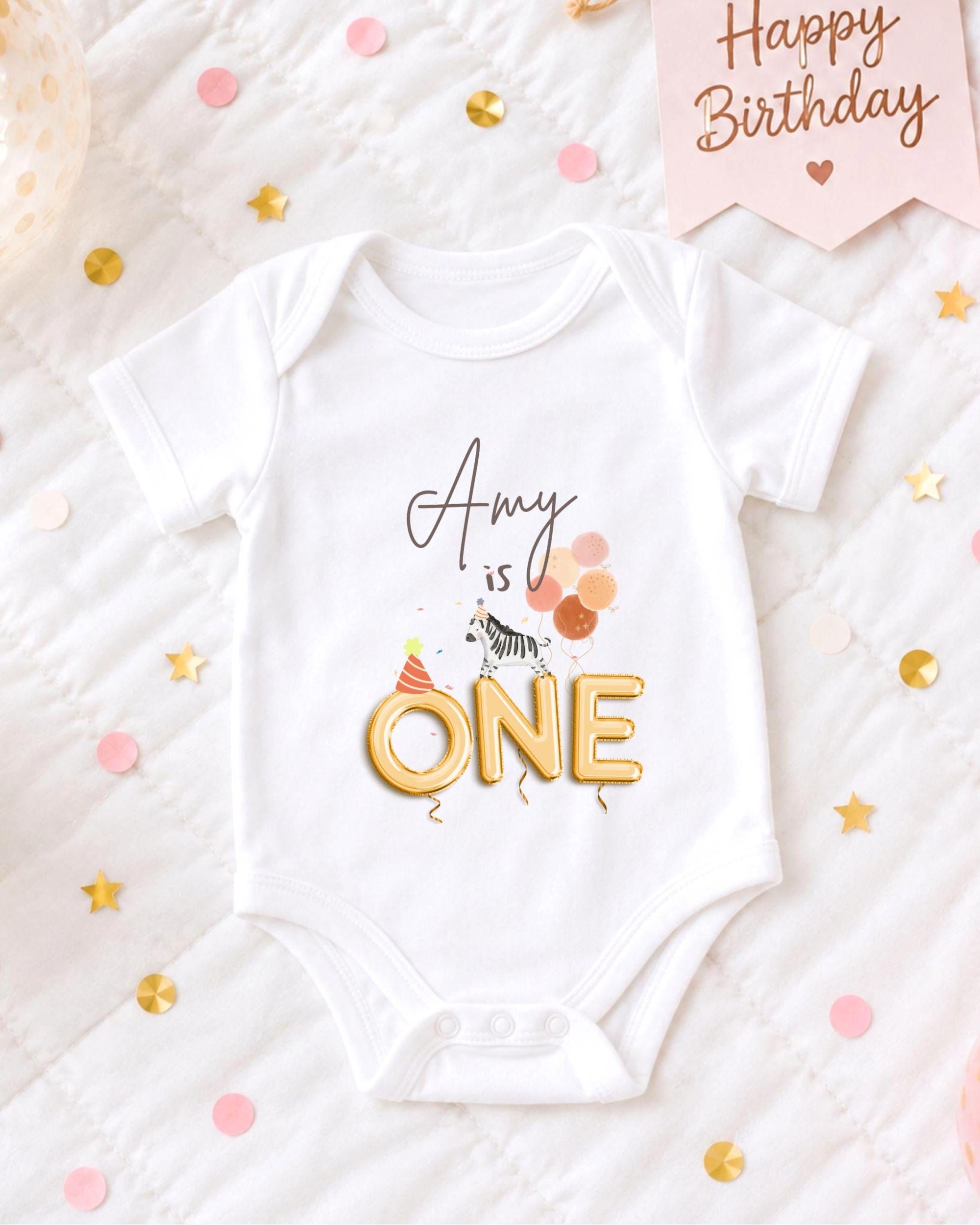 A white onesie with the text "amy is one" printed on it, surrounded by confetti and a birthday card.