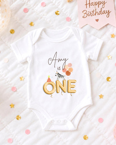 A white bodysuit with the text "amy is one" printed on it, surrounded by confetti and a birthday card.