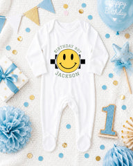 A white onesie with a smiley face and the text "Birthday Boy Jackson" printed on it, surrounded by birthday decorations and a blue balloon.