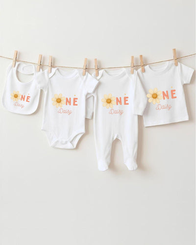 A row of four white baby clothes with the word "Daisy" printed on them, hanging on a clothesline.