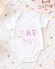 A white onesie with a pink unicorn and sprinkles, labeled "Evelyn", is displayed on a bed with confetti and a "Happy Birthday" sign.