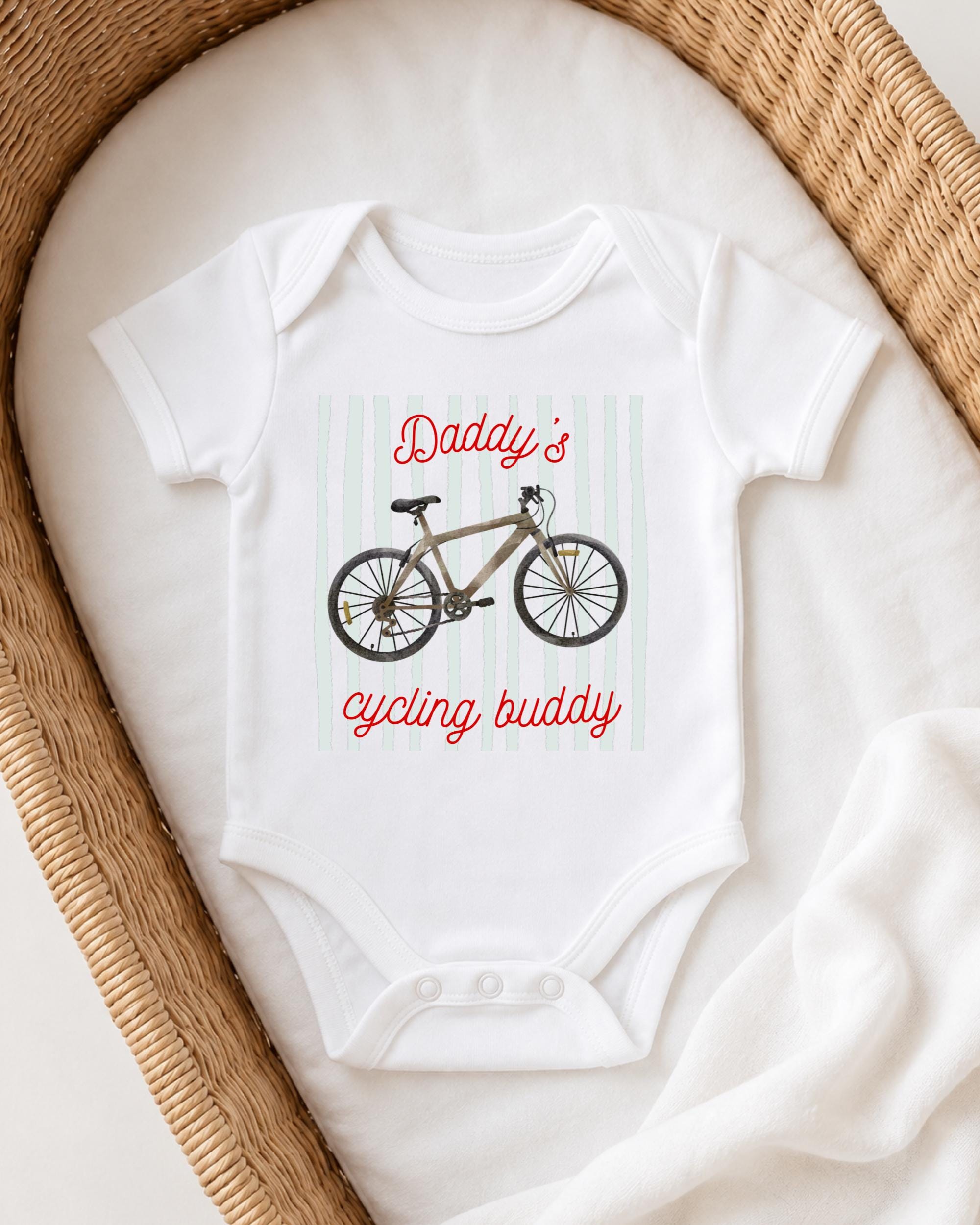 A white onesie with a bicycle graphic and the text "Daddy's cycling buddy" printed on it, placed in a woven basket.