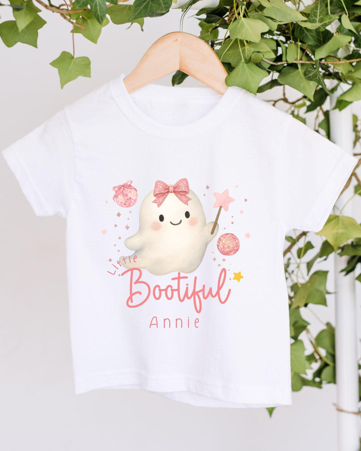 A white t-shirt with a cartoon ghost character and the text "Little Bootful Annie" printed on it, hanging on a wooden hanger against a backdrop of greenery.
