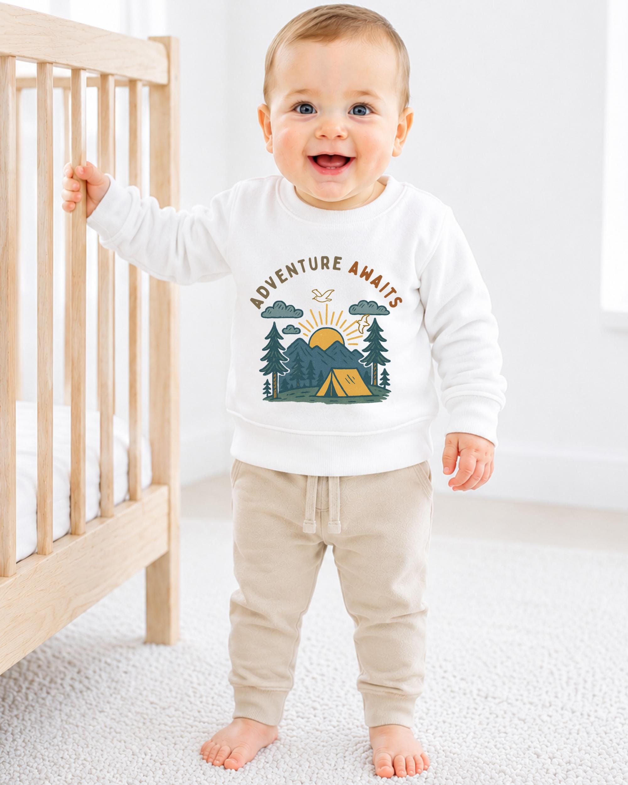 A smiling baby boy wearing a white sweatshirt with a graphic design of a tent and mountains, standing next to a wooden crib.