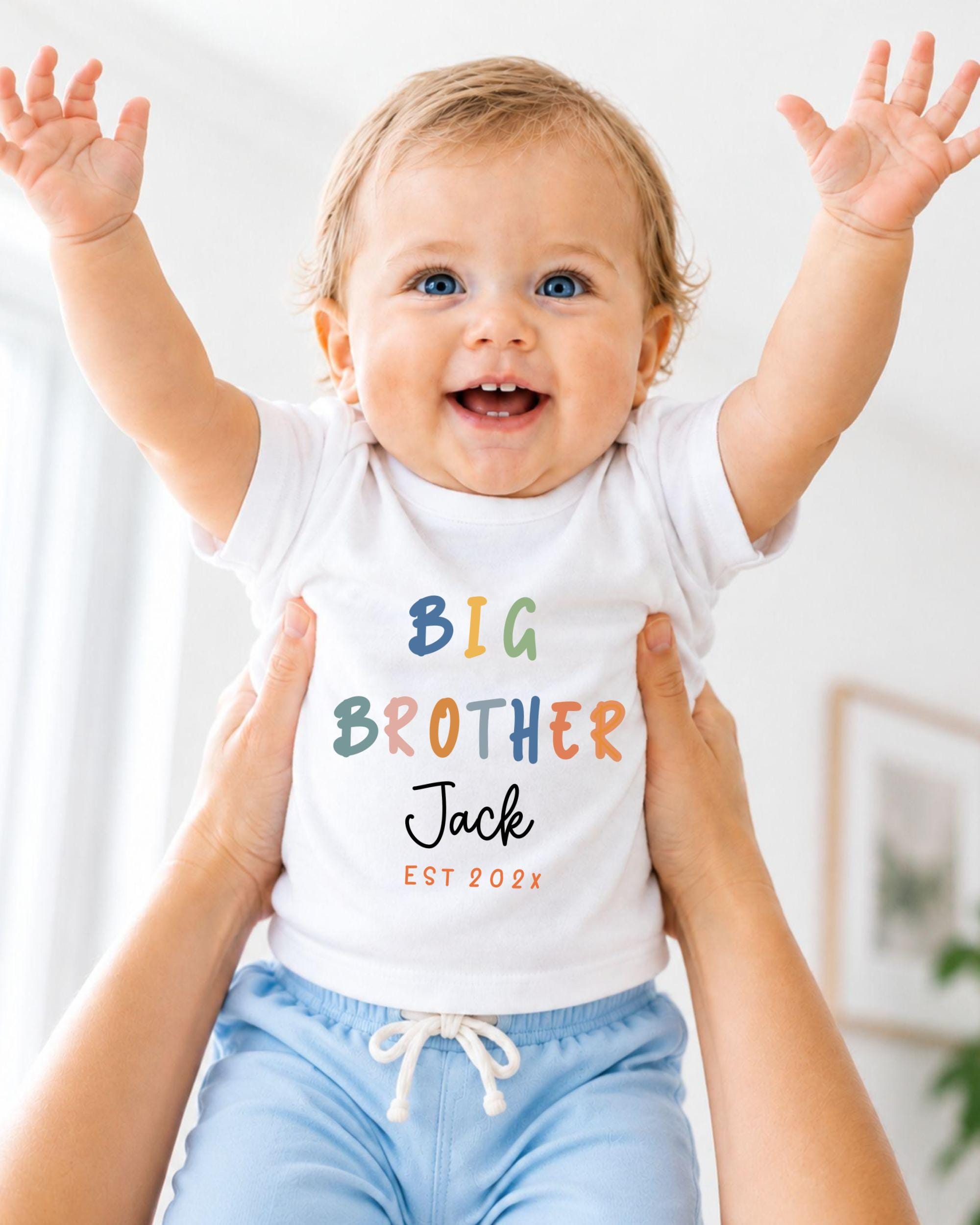 A smiling baby wearing a white shirt with the text "BIG BROTHER Jack EST 2023" is being held up by someone's arms.
