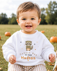 A smiling baby boy wearing a white sweatshirt with a raccoon graphic and text that says "Cute in town Hudson".