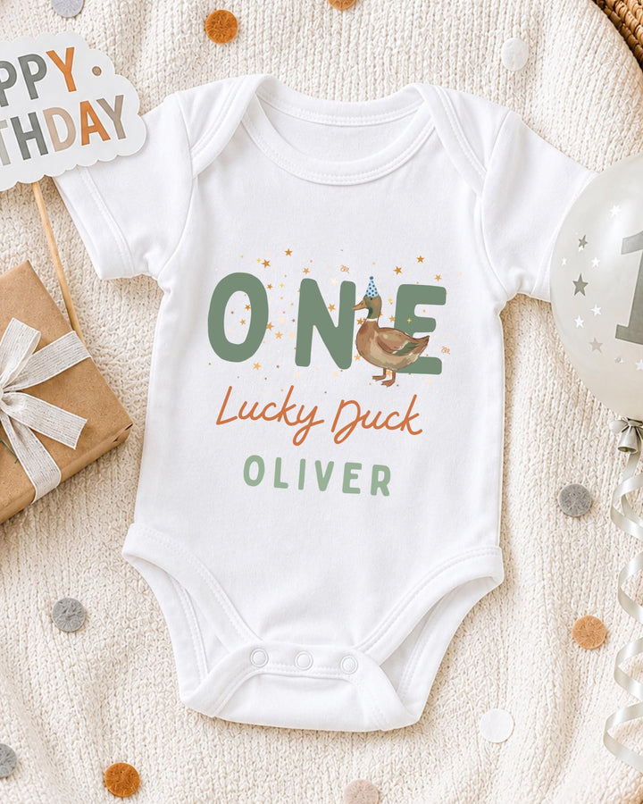 A white bodysuit with the text "ONE Lucky Duck Oliver" printed on it, along with a birthday sign and a balloon, all placed on a white blanket with confetti.