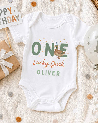 A white bodysuit with the text "ONE Lucky Duck Oliver" printed on it, along with a birthday sign and a balloon, all placed on a white blanket with confetti.
