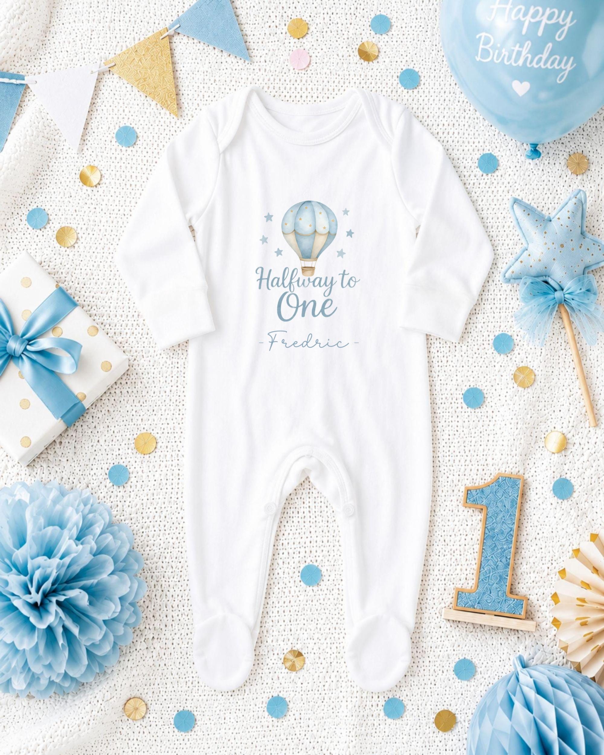 A baby onesie with the text "Halfway to One" printed on it, surrounded by baby-themed decorations such as a balloon, pom pom, and confetti.