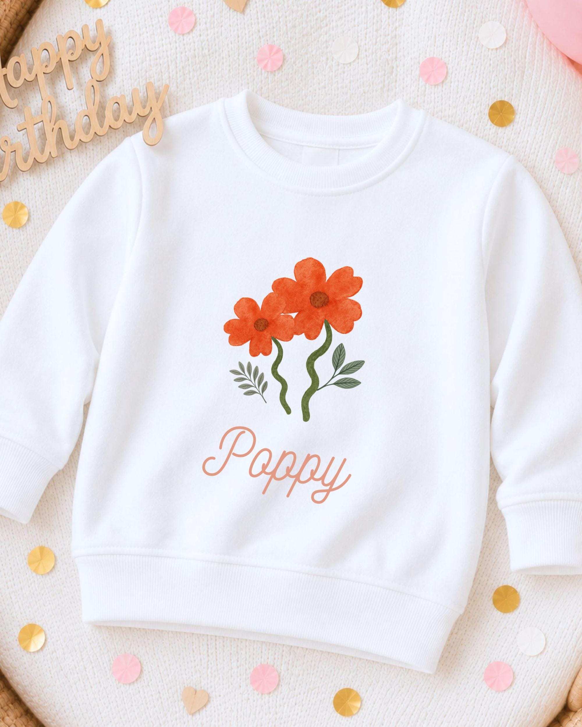 A white sweatshirt with a floral design and the name "Poppy" printed on it, surrounded by confetti.