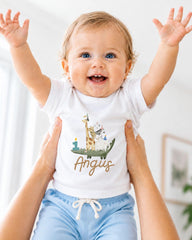 A smiling baby wearing a white t-shirt with a giraffe and the name "Angus" printed on it, being held up by someone's arms.